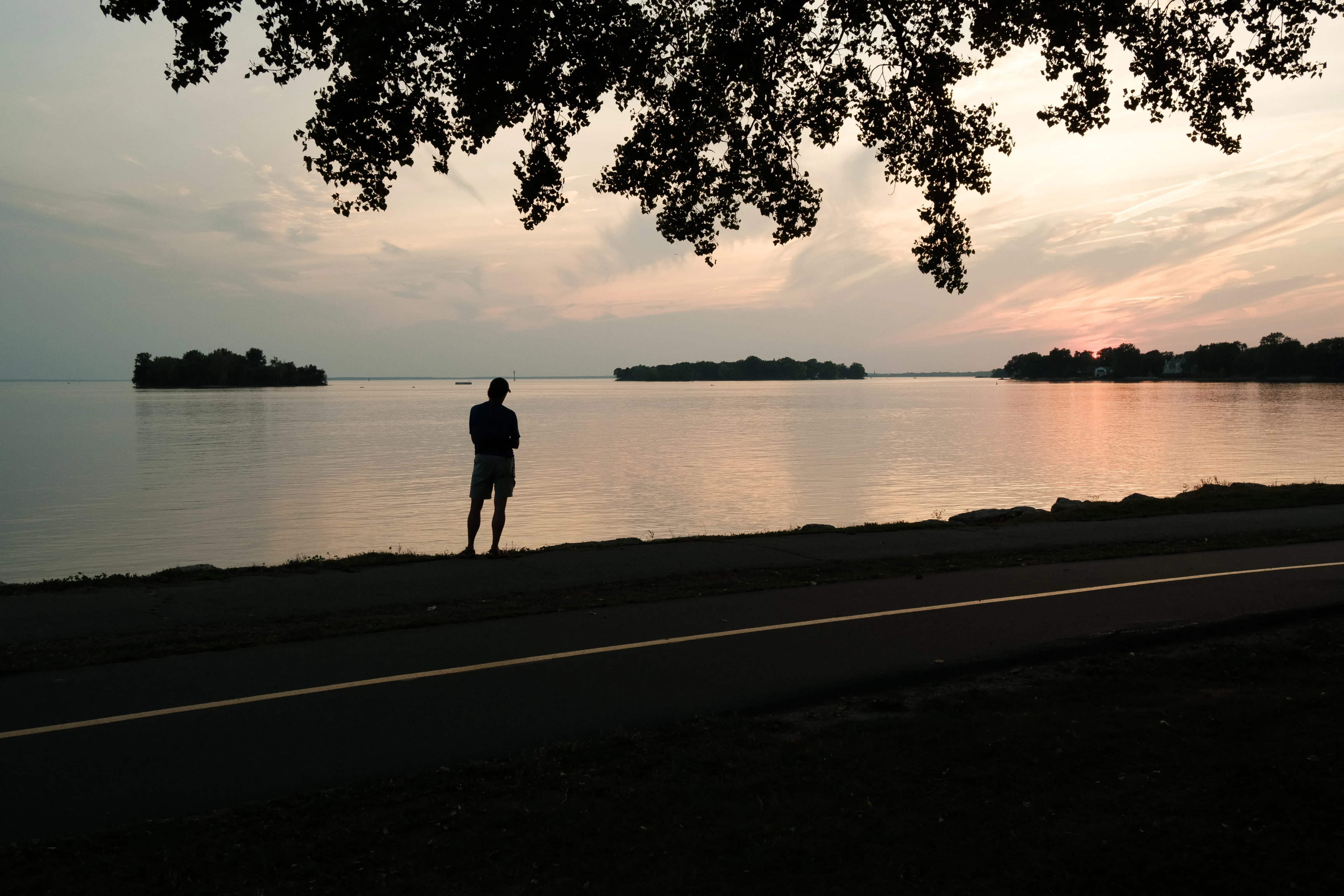 a man standing on the side of a road next to a body of water.The sky is a gradient of pink and blue, and the water reflects the colors.