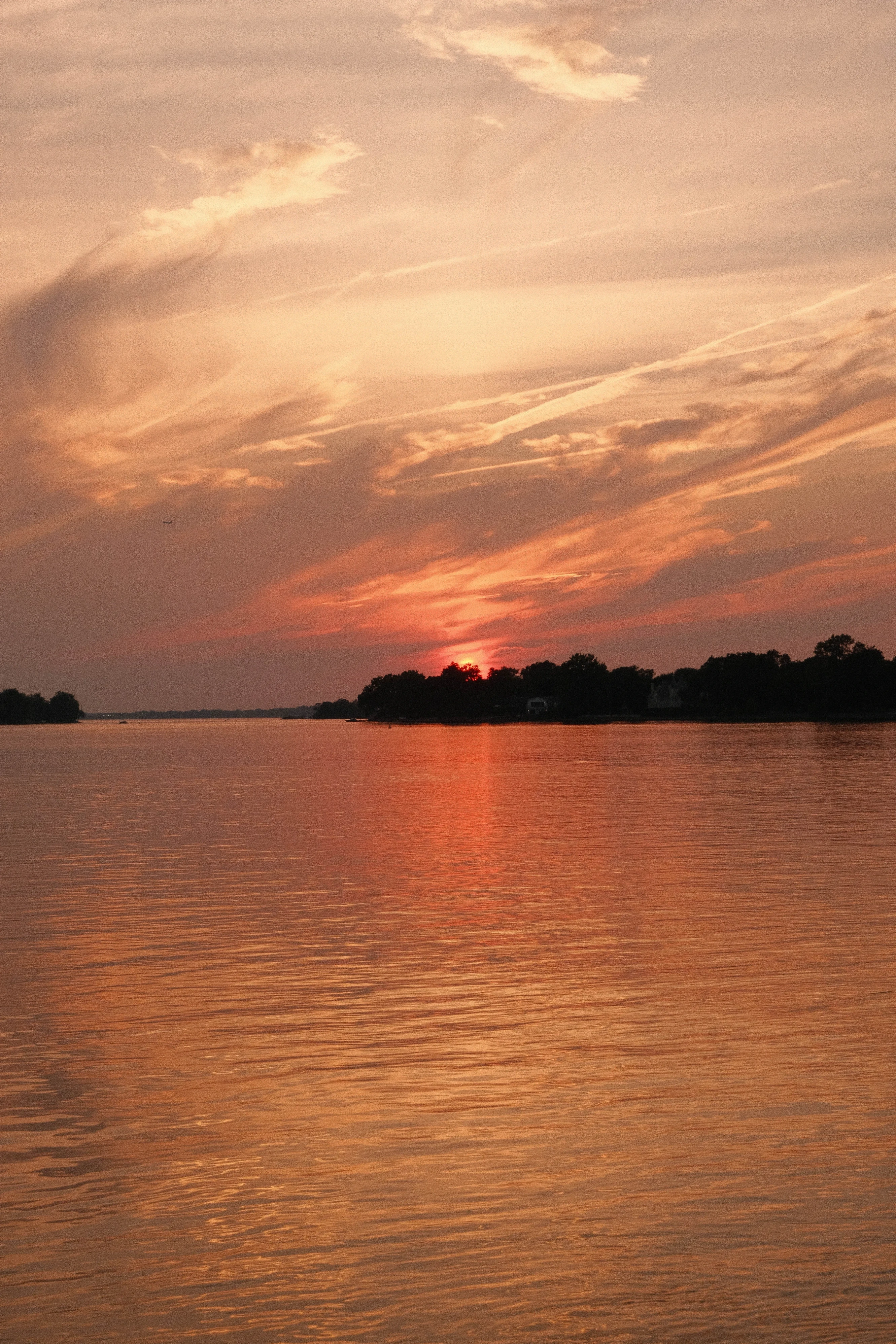 A beautiful sunset over a calm lake. The sky is ablaze with orange and pink hues, with wispy clouds streaking across it. The sun dips below the horizon, casting a warm glow on the water. Silhouetted trees line the shore.