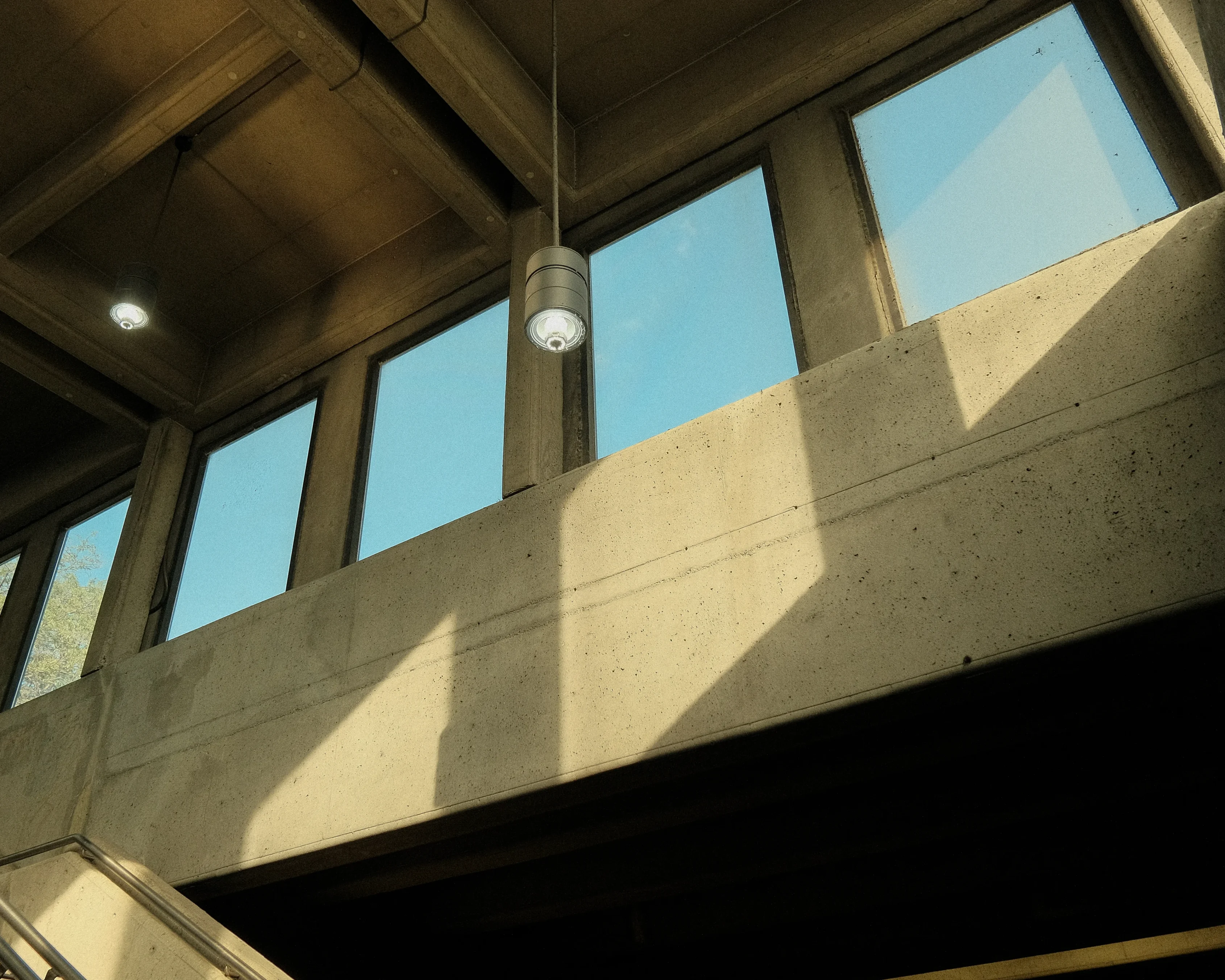 A photo of a modern building interior with a concrete ceiling and a row of windows. Sunlight streams through the windows, casting shadows on the walls and floor. A hanging lamp is visible in the center of the frame.