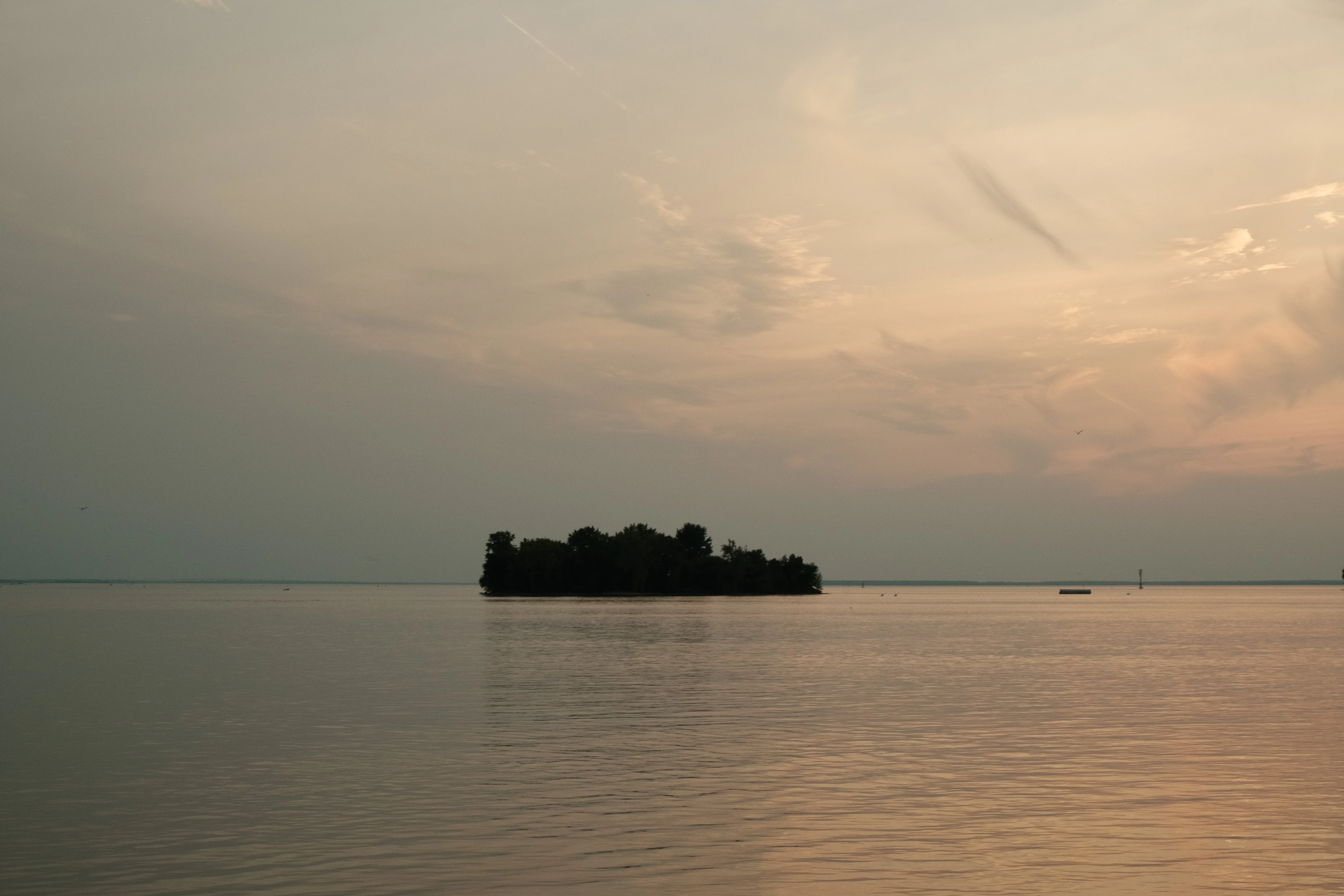 A small, forested island sits in the middle of a calm lake at sunset. The sky is a soft pink and blue, and the water is still.