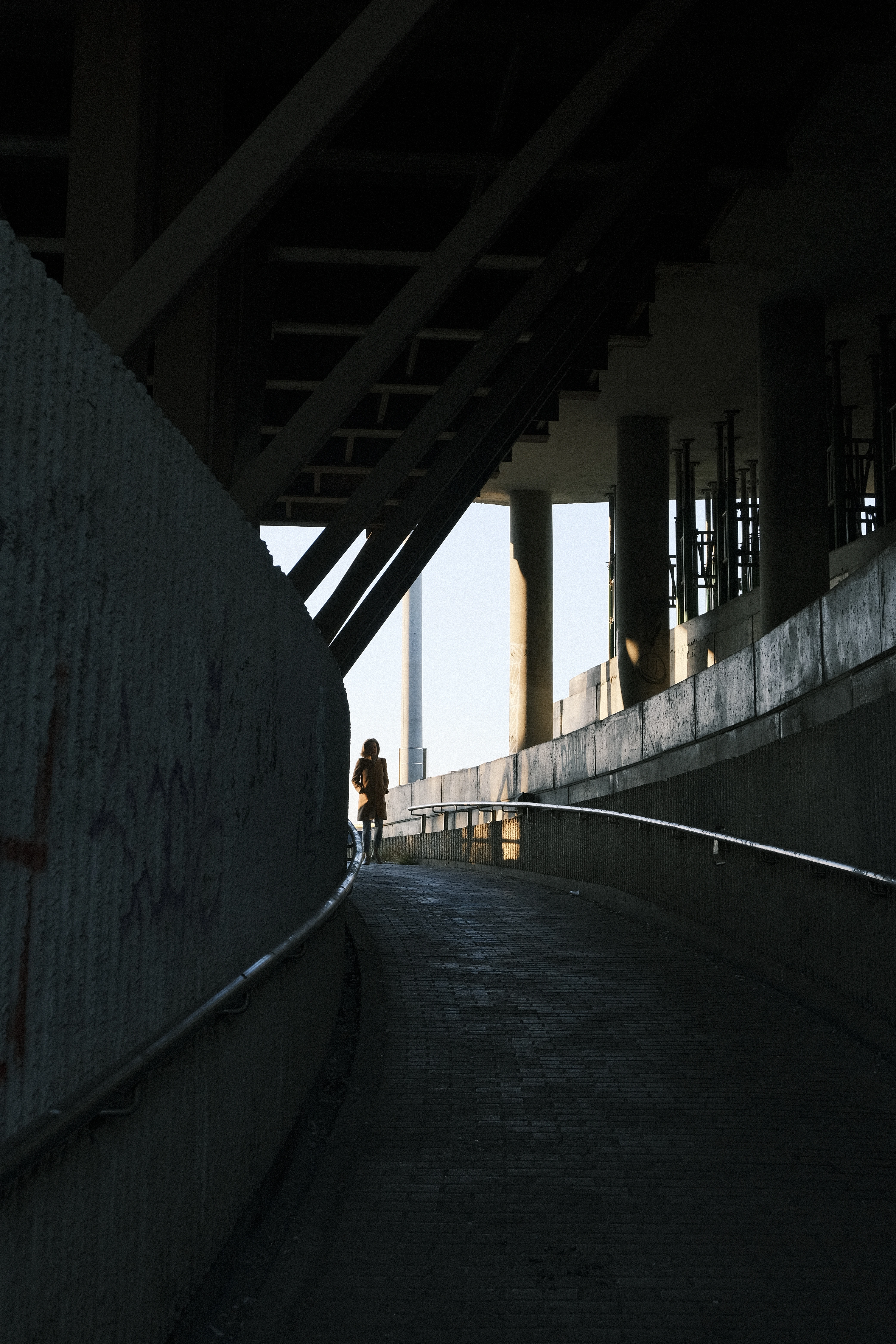  A photo of a person walking alone on a ramp under a bridge. The person is wearing a dark coat and is walking away from the camera. The underside of the bridge is dark and has metal beams and supports. There is graffiti on the wall to the left.