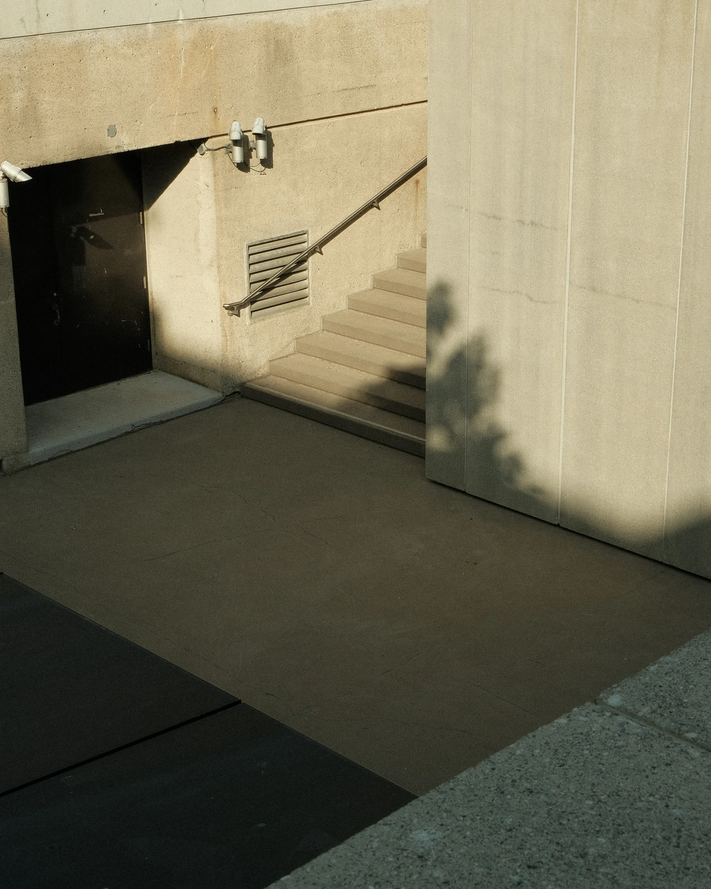 A concrete staircase leads down to a dark doorway. The stairs are partially in shadow, and there is a vent and a handrail on the side.