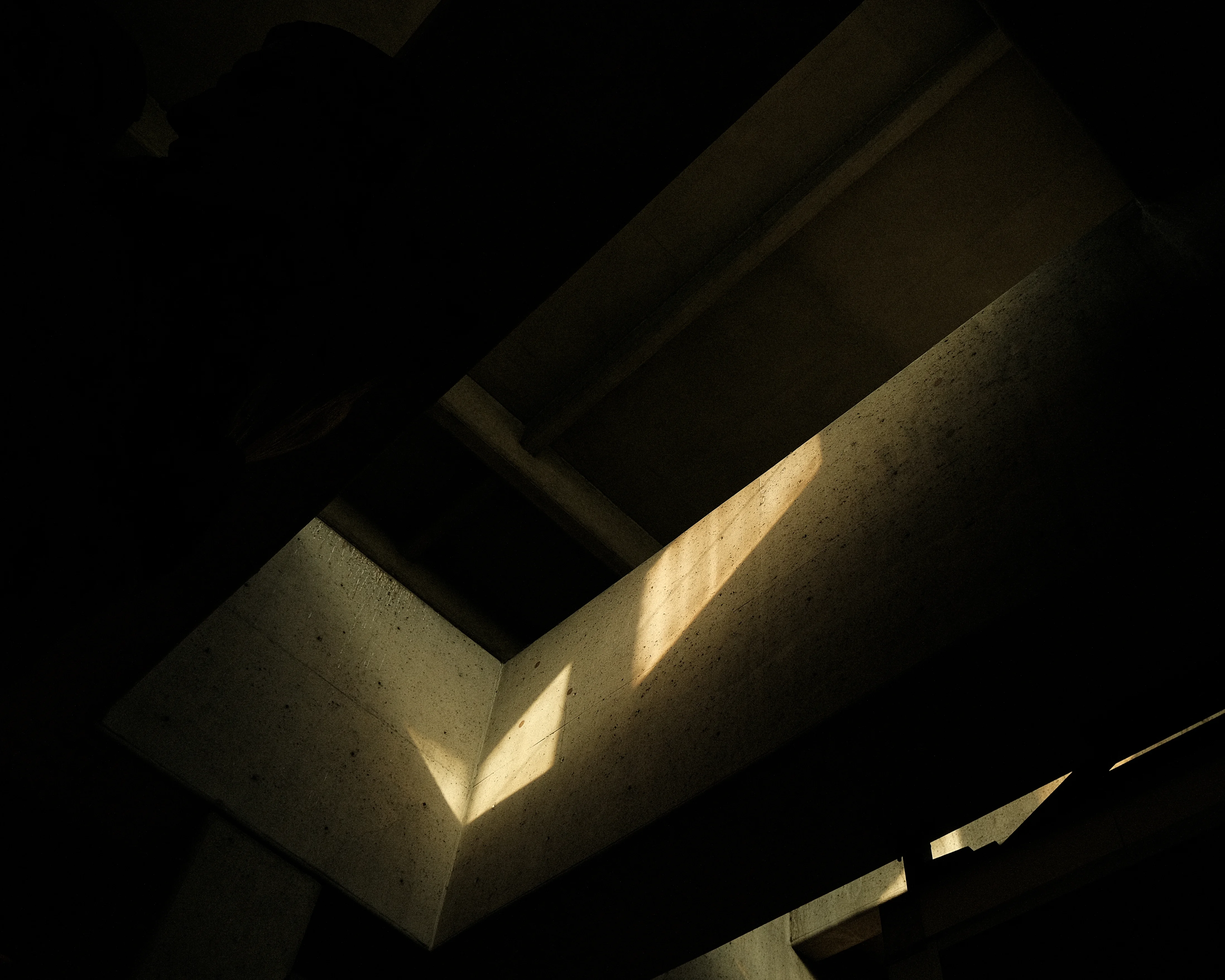 A narrow beam of light shines through a crack in a dark, concrete ceiling. The light illuminates a small section of the ceiling and casts a shadow on the wall below.