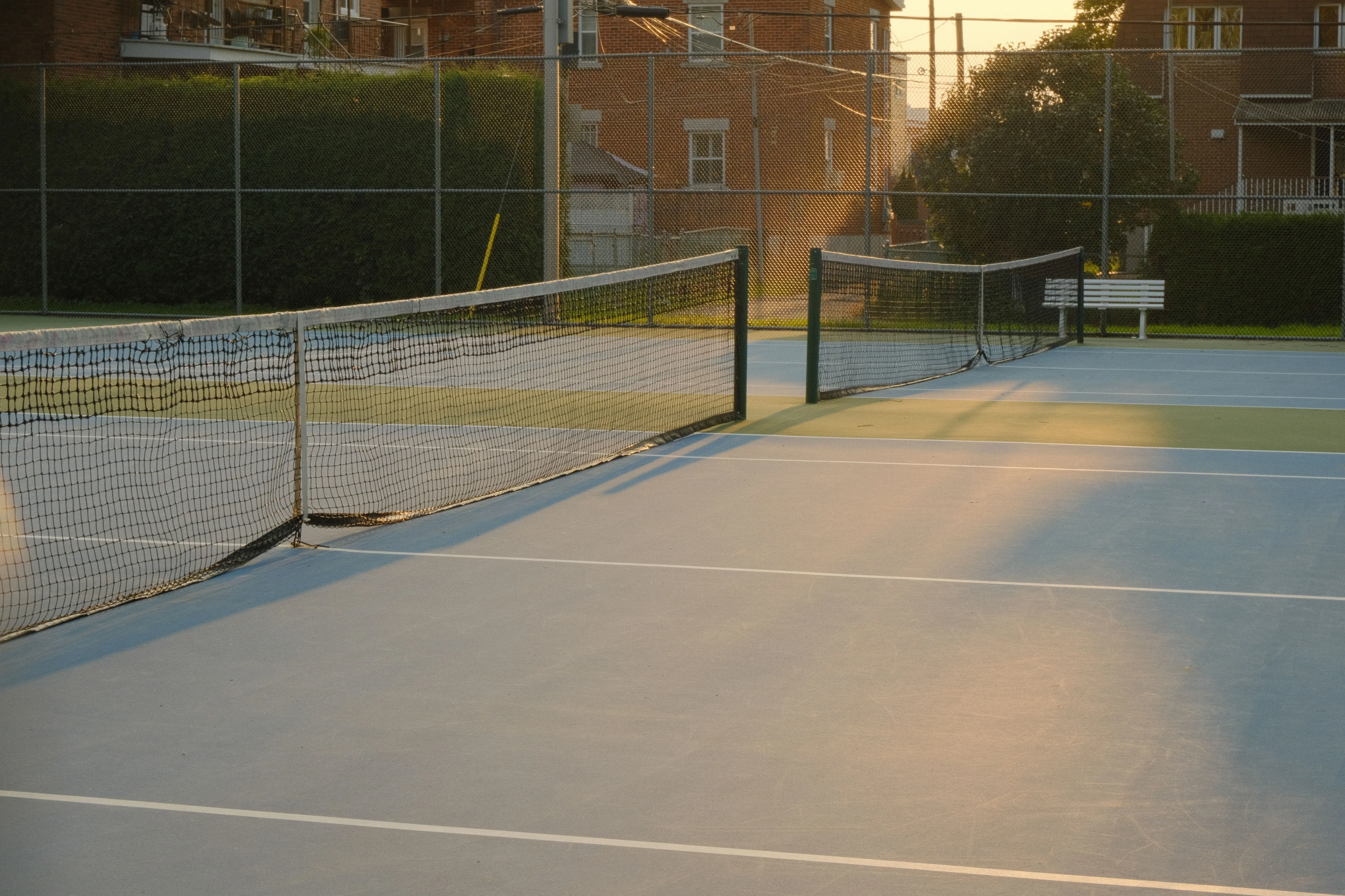 A tennis court with a blue surface and white lines. Two nets divide the court in half. The sun is setting, casting a warm glow on the court.