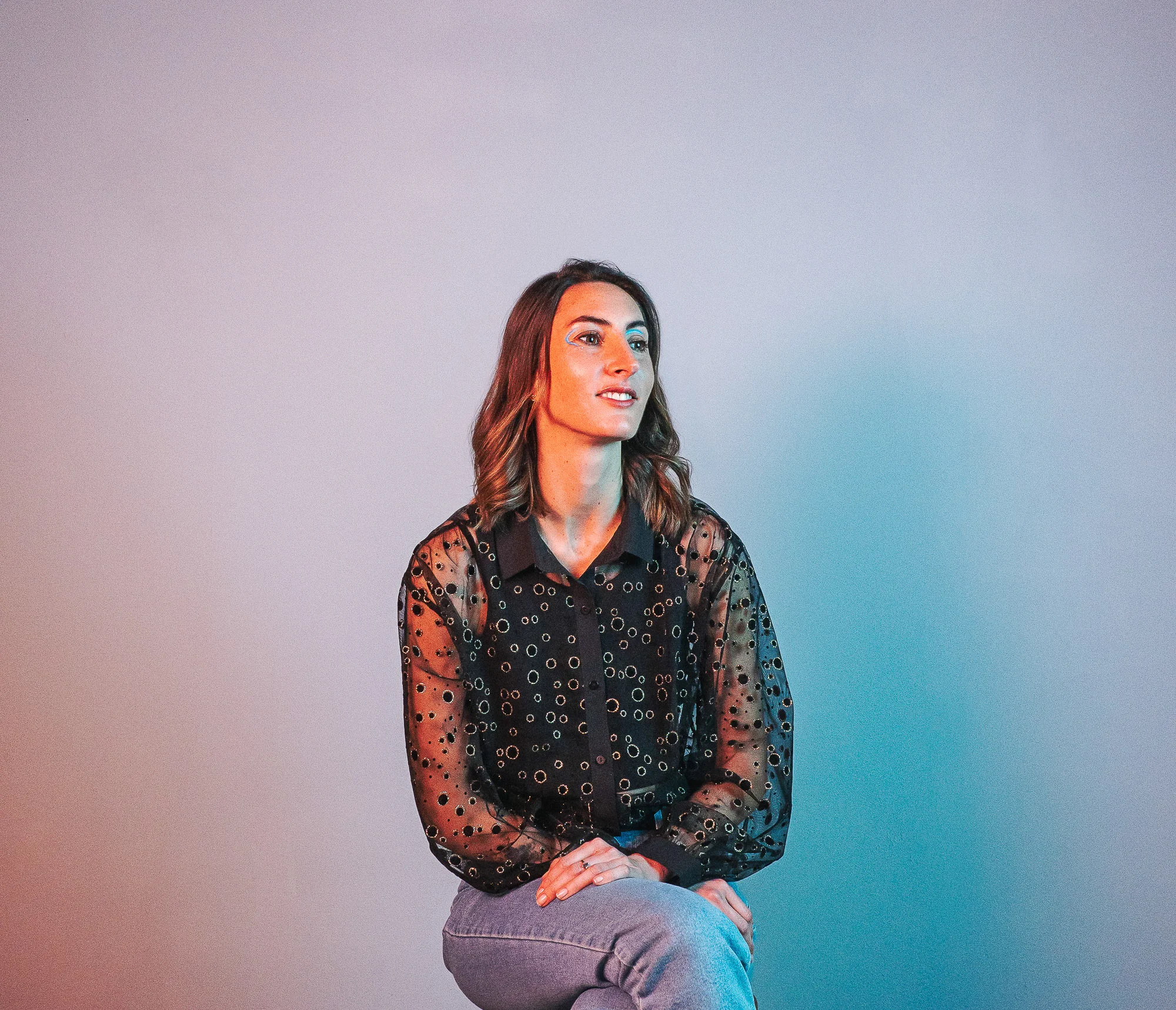 A person with long brown hair sits on a stool, wearing a sheer polka dot shirt and jeans against a gradient background.