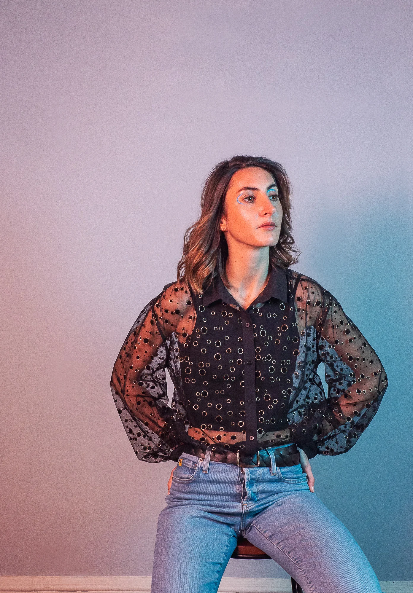 A person with shoulder-length hair sits on a stool, wearing a sheer black patterned blouse and jeans, against a light grey background.